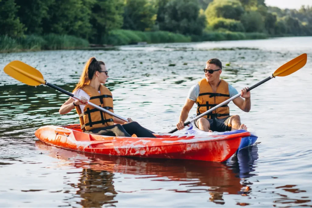 Two people in orange kayaks paddle together on a tranquil river, wearing life jackets and enjoying a sunny day.