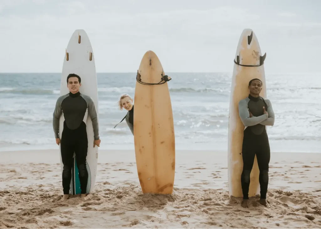 Three surfers in wetsuits stand on a beach, two holding surfboards, while one playfully peeks from behind a board.