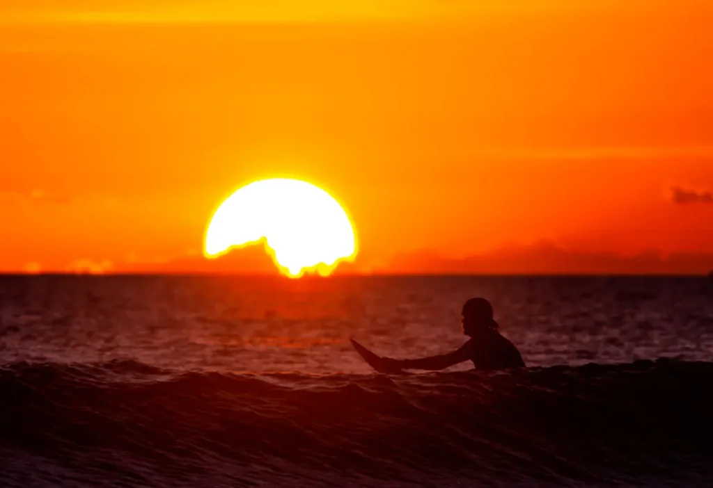 Silhouetted surfer catching waves against a vibrant sunset backdrop over the ocean horizon.