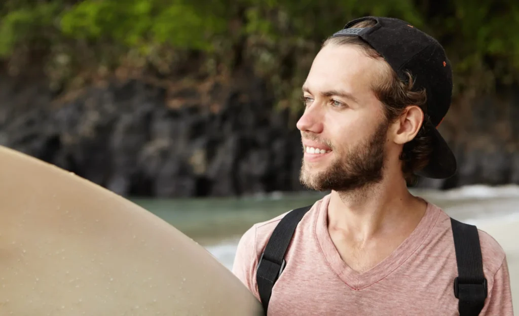 Smiling surfer holding a surfboard, standing on the beach with lush greenery in the background.