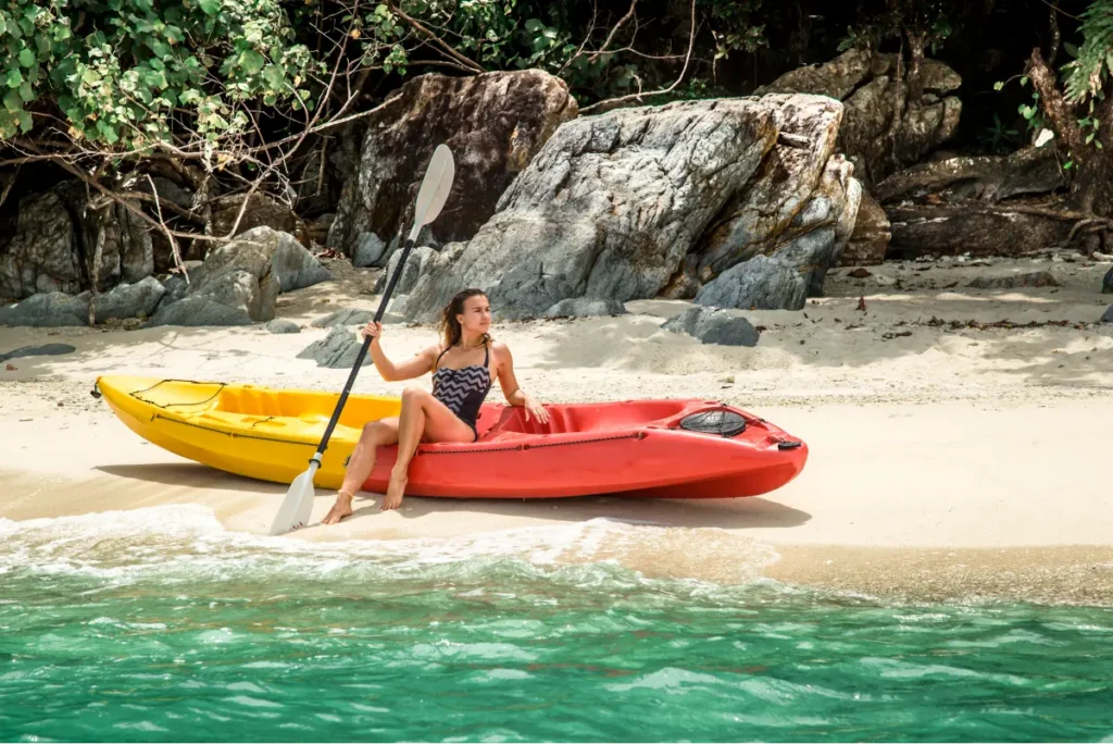 A woman sits in a kayak on a sandy beach, holding a paddle, surrounded by lush greenery and rocks.